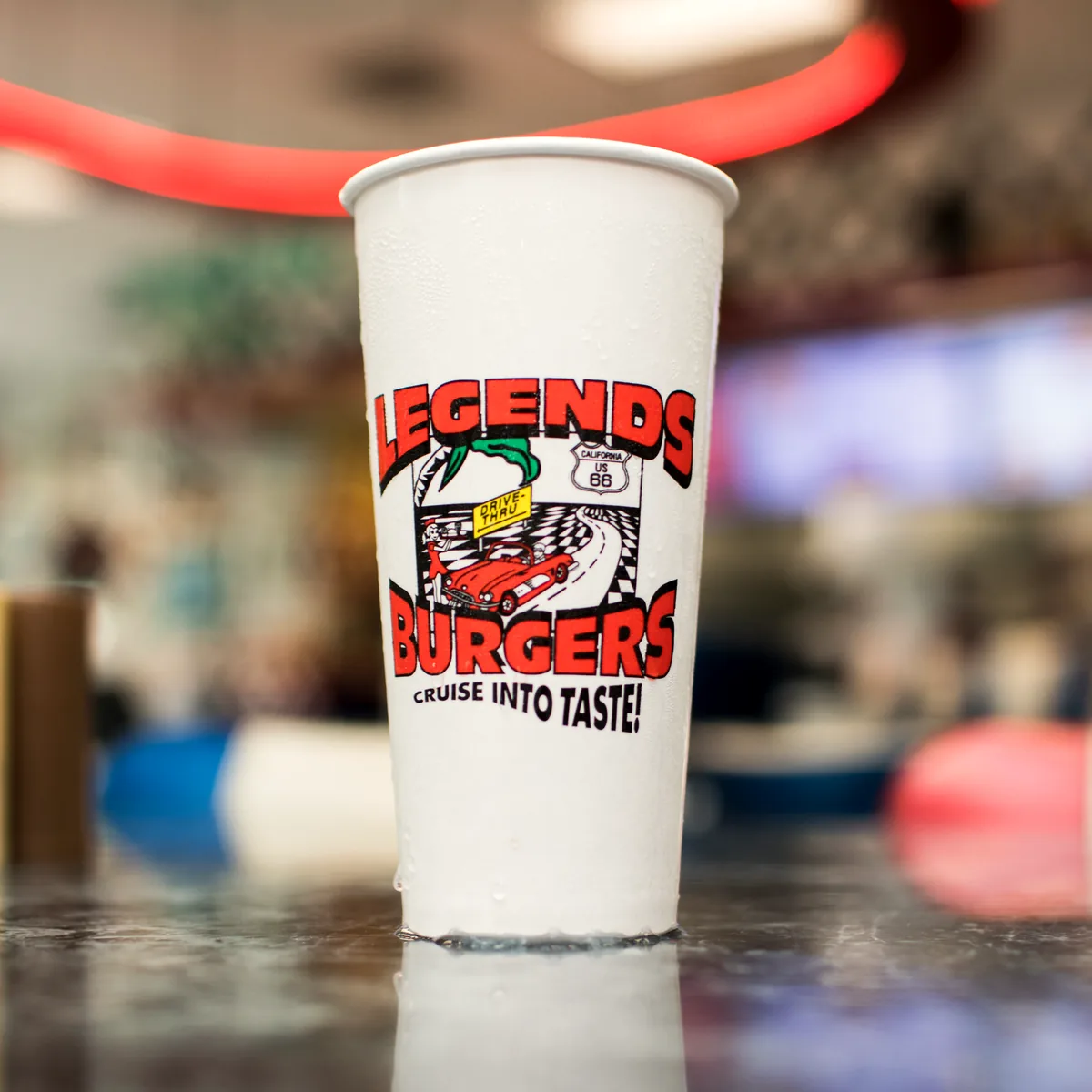 A Legends Burgers paper cup on the counter, printed with the red script
                      logo, the 1955 Chevrolet and the words &lsquo;Cruise into taste!&rsquo;,
                      with the dining room out of focus behind it