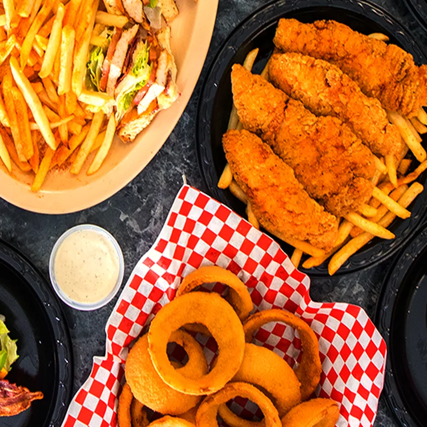 Breaded chicken strips and fries on a black plate, with a basket of onion rings and a cup of ranch